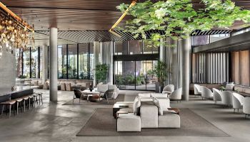 A spacious, modern lobby features elegant seating, hanging lights, large windows, and greenery, creating a welcoming atmosphere.
