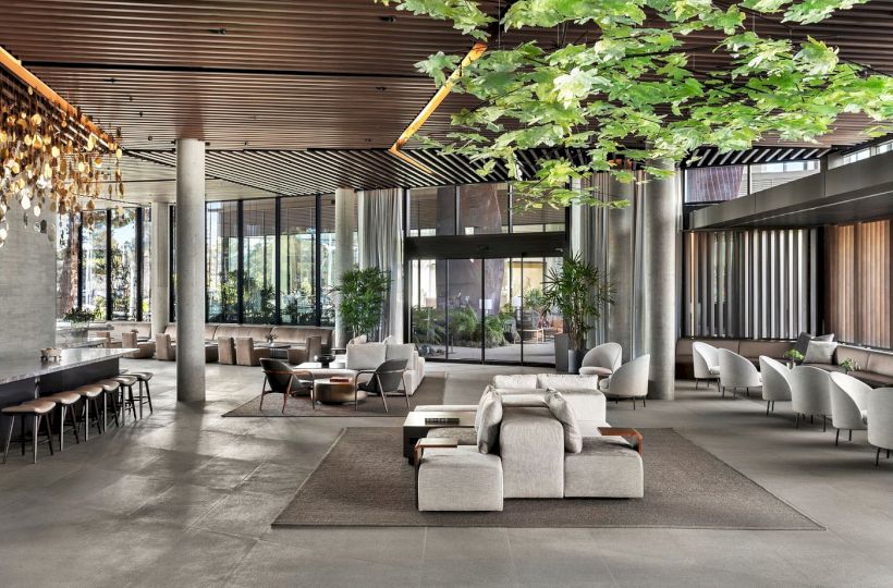 A spacious, modern lobby features elegant seating, hanging lights, large windows, and greenery, creating a welcoming atmosphere.