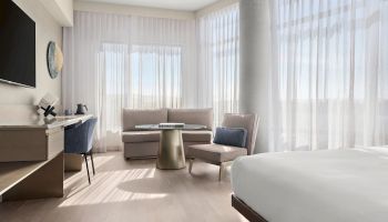 A modern hotel room with a bed, desk, chair, TV, and a sitting area by large windows with sheer curtains.