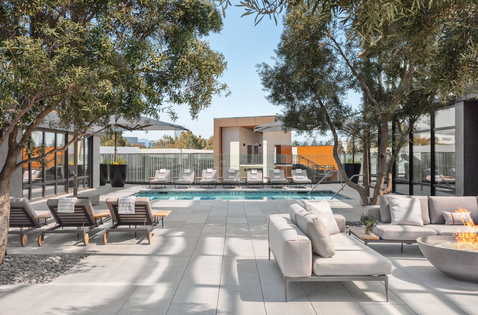 A modern outdoor patio with trees, loungers, sofas, a fire pit, and a pool, surrounded by glass walls and buildings in the background.