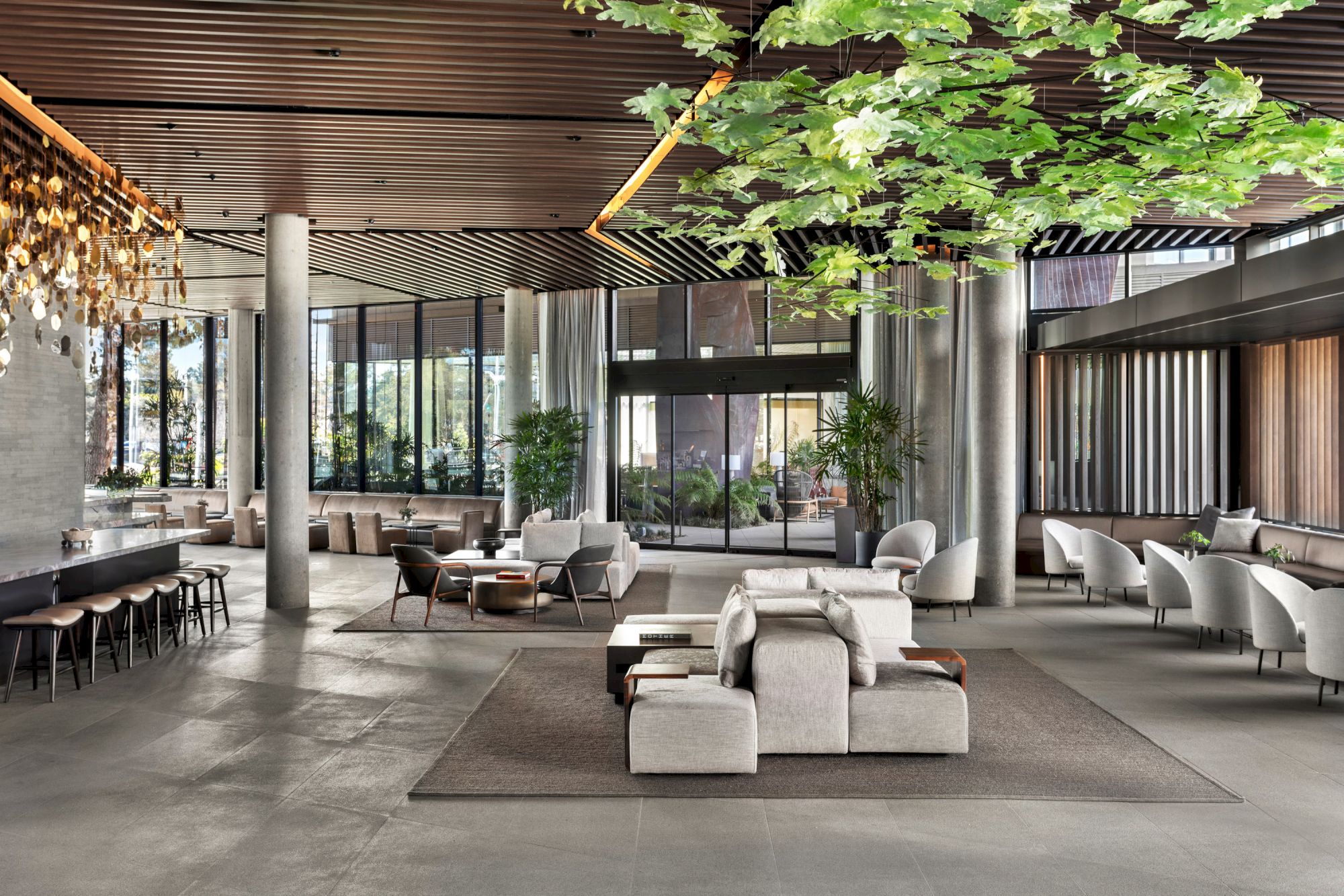 A modern lobby with sleek furniture, large windows, greenery on the ceiling, and a long, elegant chandelier above the seating area.
