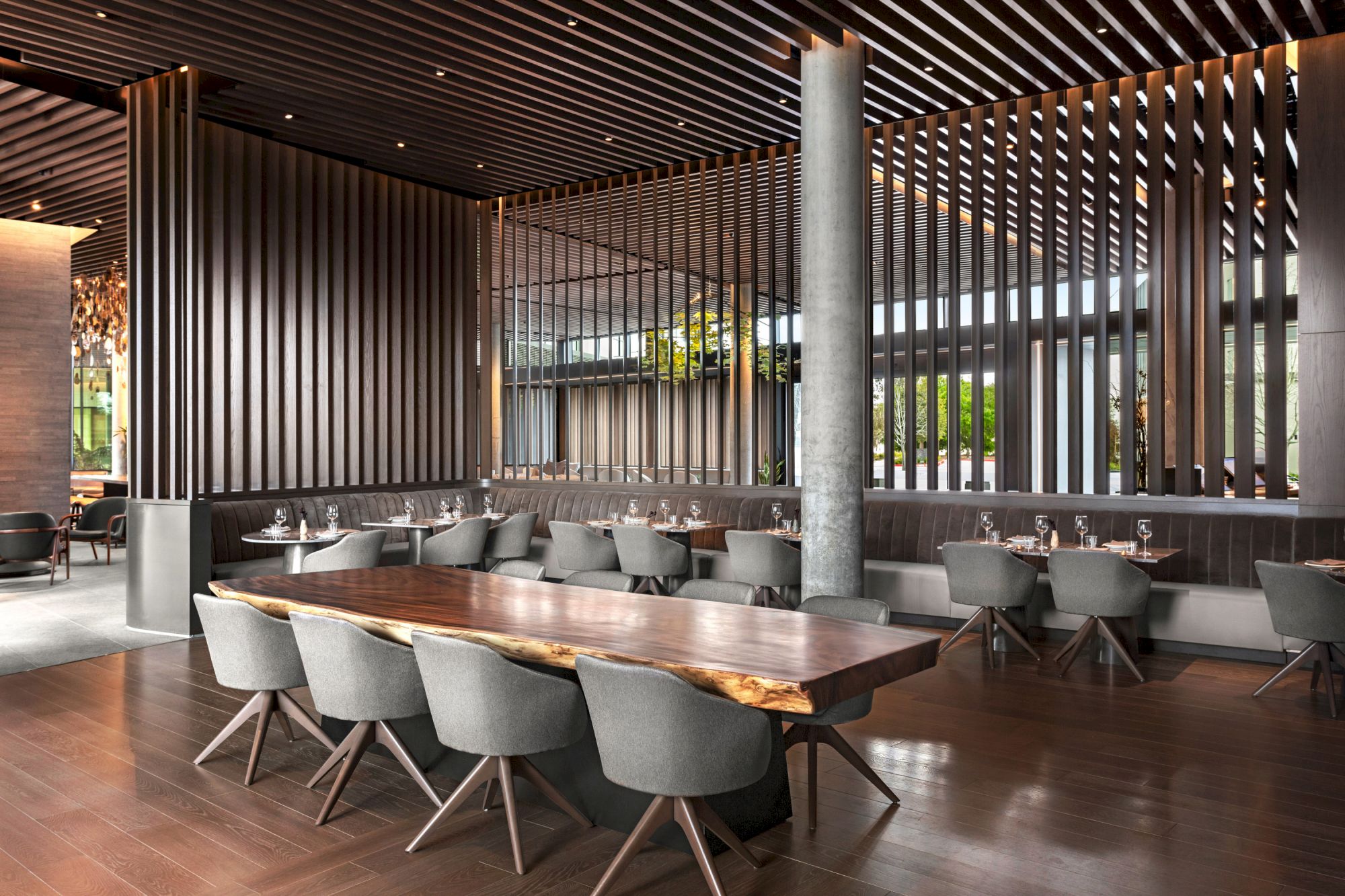 The image shows a modern restaurant interior with a large wooden table, gray chairs, and vertical wooden slats on the walls for decor.