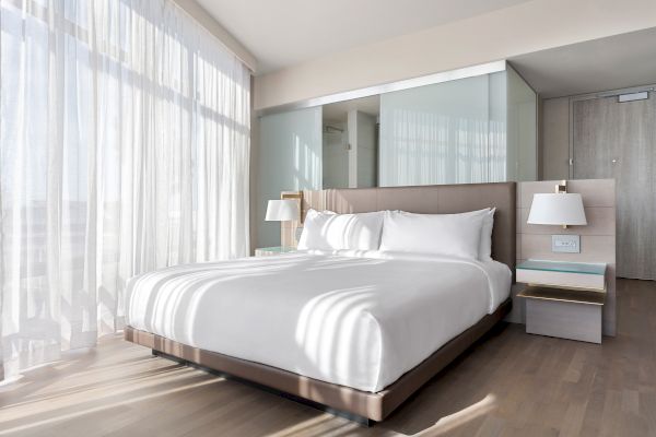 A bright hotel room with a large bed, bedside tables, lamps, and sunlight streaming through sheer curtains.