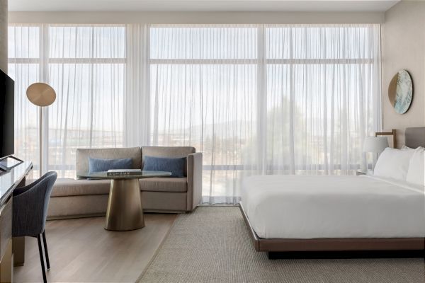 A modern hotel room with a cozy sofa, bed, desk, and lamp, featuring large windows with sheer curtains providing a bright, airy feel.