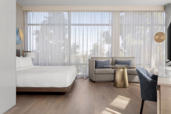 A modern hotel room with a bed, sofa, round table, and large windows with sheer curtains letting in natural light.