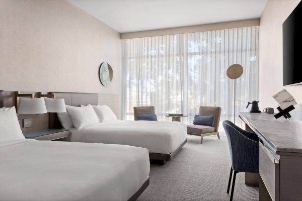 A modern hotel room with two beds, a desk, chair, and a sitting area by the window with sheer curtains and gentle lighting.