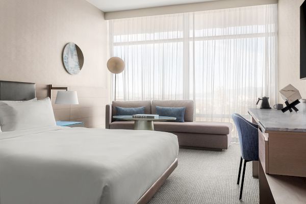 A modern hotel room with a bed, sofa, desk, and a large window with sheer curtains, creating a bright and cozy atmosphere.