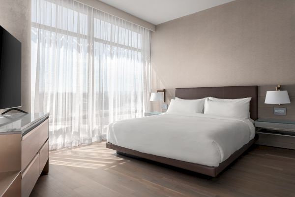 A modern hotel room with a large bed, white linens, soft lighting, and a TV, featuring sheer curtains on tall windows enhancing the space.