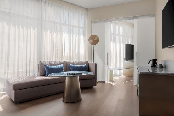 A modern living room with a sofa, blue pillows, round glass table, TV, sliding doors, sheer curtains, and a kettle on a counter.