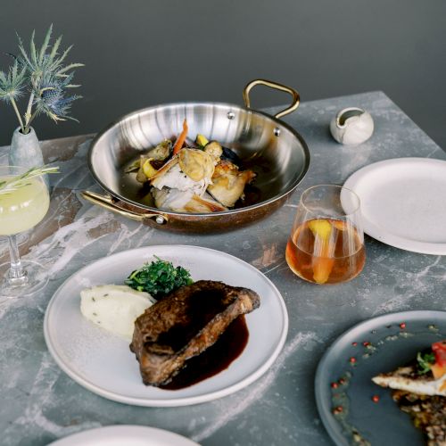 A table with various dishes including meat, fish, and vegetables, alongside drinks and an empty plate.