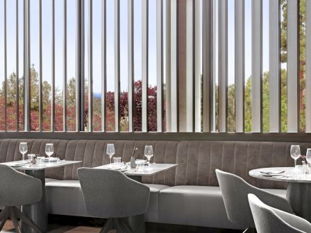 The image shows a modern restaurant interior with sleek gray chairs, tables, and large vertical windows overlooking trees outside.