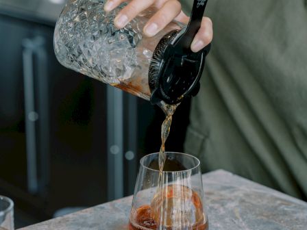 A person pours a drink from a crystal mixing glass with a strainer into a glass with an ice sphere on a marble table.