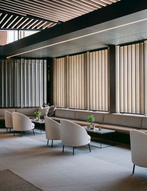 A modern lounge area with beige chairs, tables, plants, and large windows with vertical blinds.