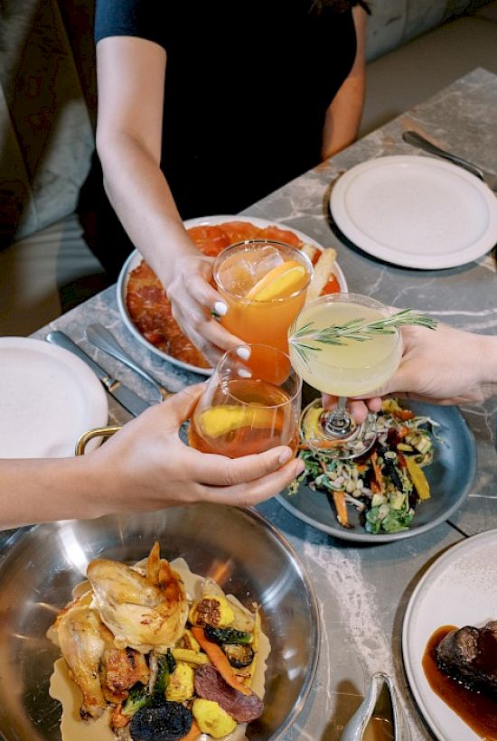 A group of people clinking their drinks over a table filled with a variety of delicious dishes in a restaurant setting.