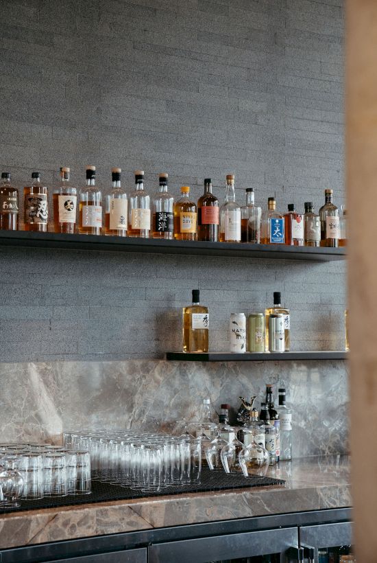 The image shows shelves with various bottles, possibly alcohol, and bar equipment, including glasses and dispensers, on a textured background.