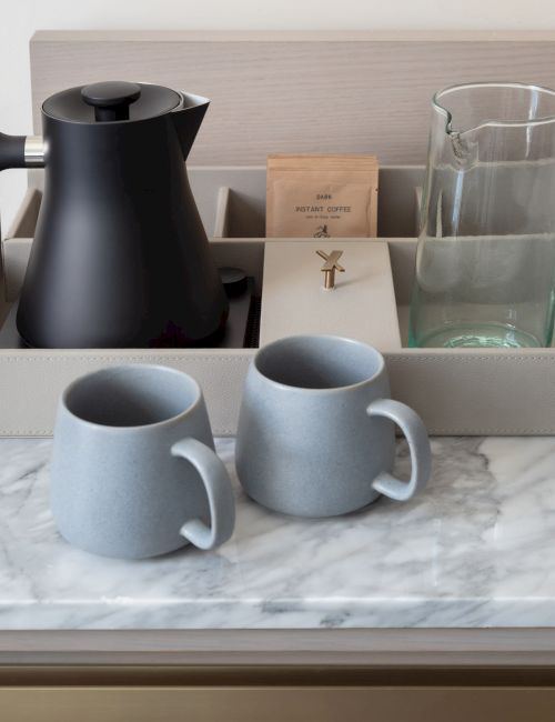 A coffee or tea setup on a marble countertop, with a black kettle, glasses, cups, and a small selection of tea or coffee items in a tray.