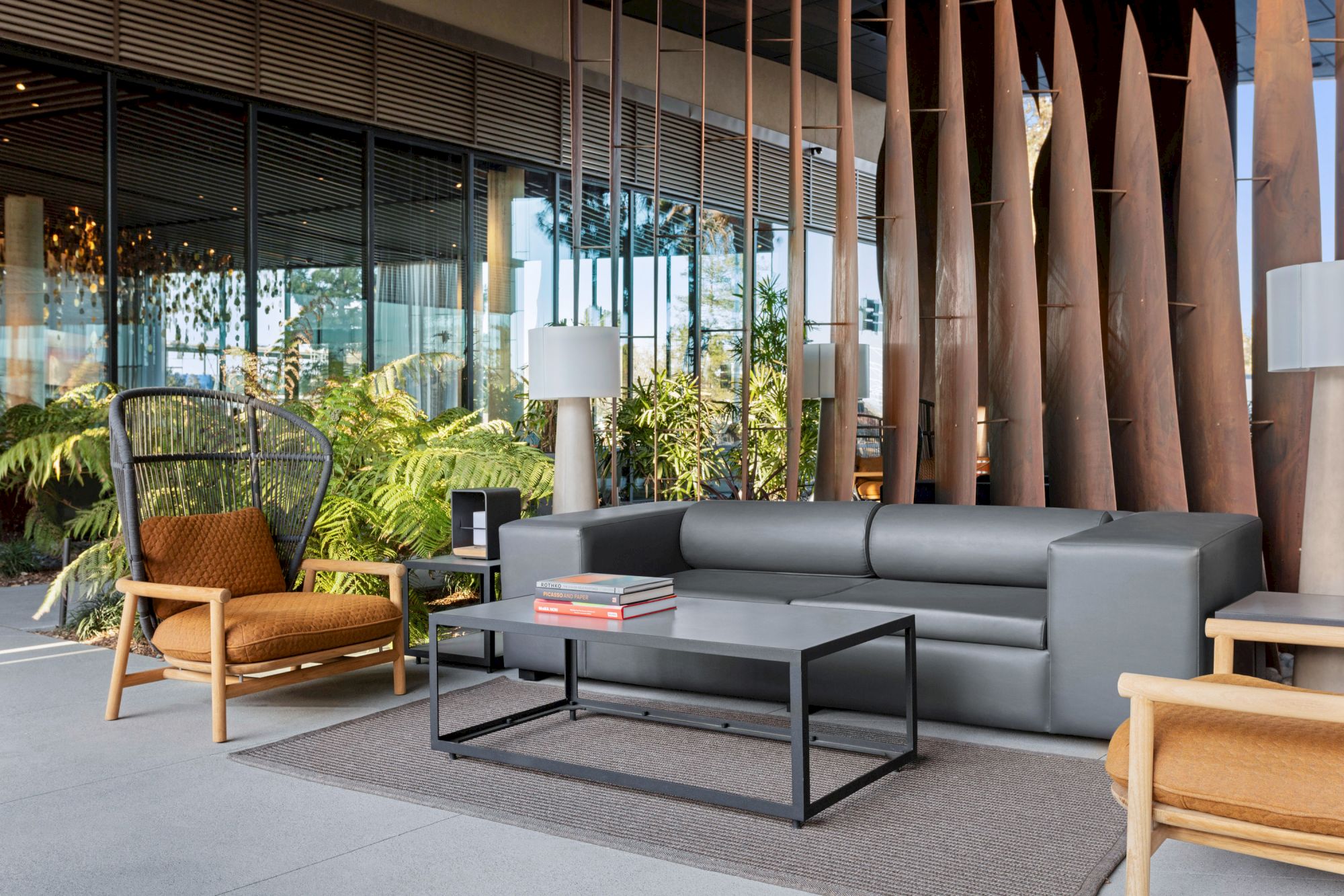 The image shows a modern patio with a gray sofa, chairs, table, books, tall decorative panels, and lush greenery in the background.