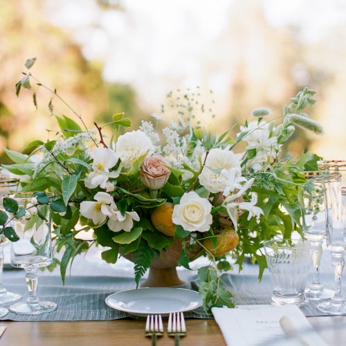 A floral centerpiece with white and pink flowers, greenery, surrounded by glassware on a table setting outdoors.