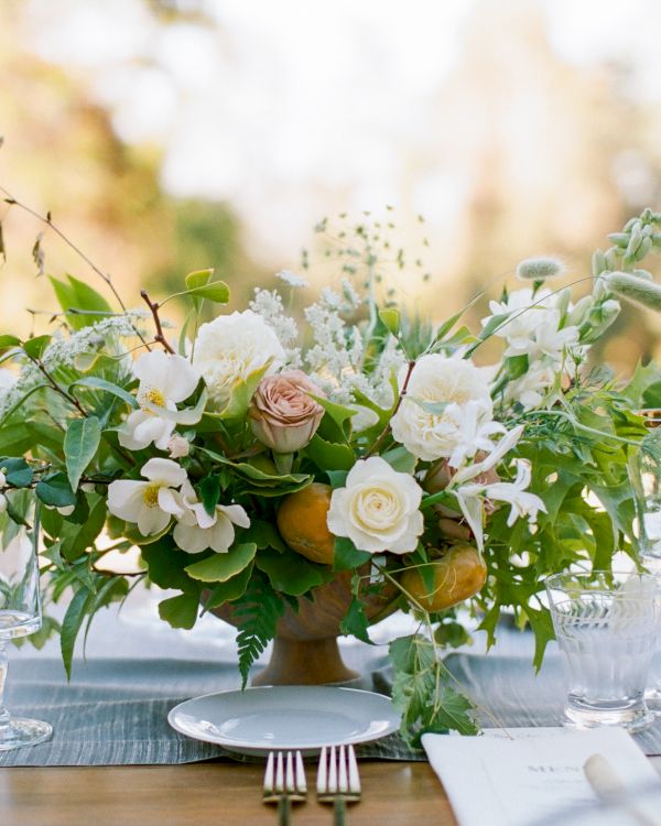 A floral centerpiece with white and pink flowers, greenery, surrounded by glassware on a table setting outdoors.