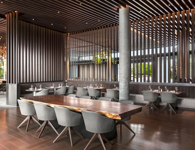 The image depicts a modern, stylish dining area with a long wooden table, gray chairs, and a wooden slat interior design.