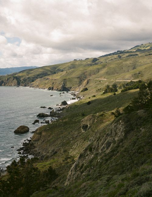A coastal landscape features rolling hills, rocky cliffs, a blue ocean, and a partly cloudy sky, creating a serene and picturesque view.