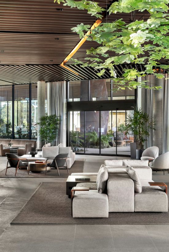 Modern lobby with plush seating, indoor plants, large windows, and a stylish ceiling with leaf decorations.