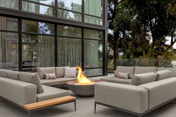 A modern outdoor seating area with gray sofas surrounds a lit fire pit, positioned near a building with large glass windows.