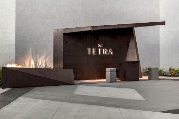 A modern entrance with "TETRA" displayed on a dark panel, flame feature to the left, and a minimalist design in a gray setting.