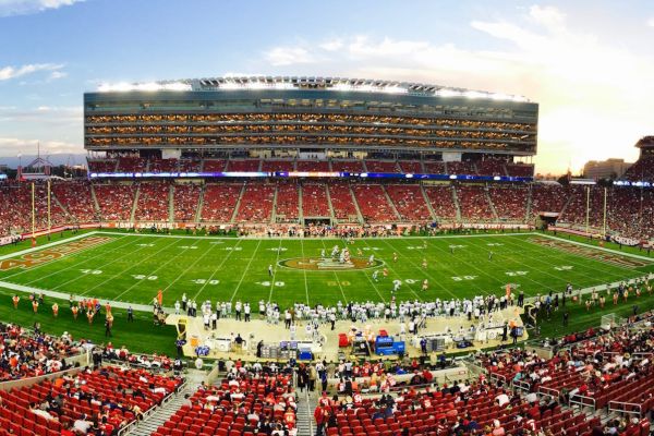 A large football stadium filled with spectators, teams on the field, scoreboard visible, all under a partially cloudy sky during sunset.