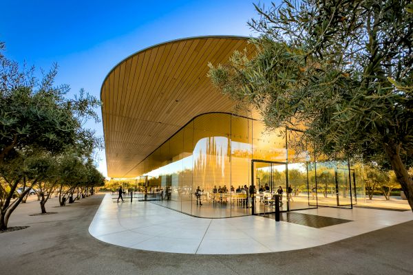 A modern building with curved glass walls and a sleek wooden roof, surrounded by trees and people gathered around it.