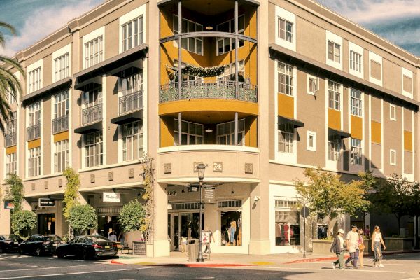 A multi-story building with shops at street level, located on a sunny corner with people walking by and cars parked nearby.