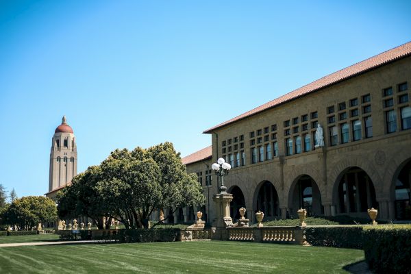A grassy lawn leads to a large building with arched windows and a red-roofed tower against a clear blue sky.