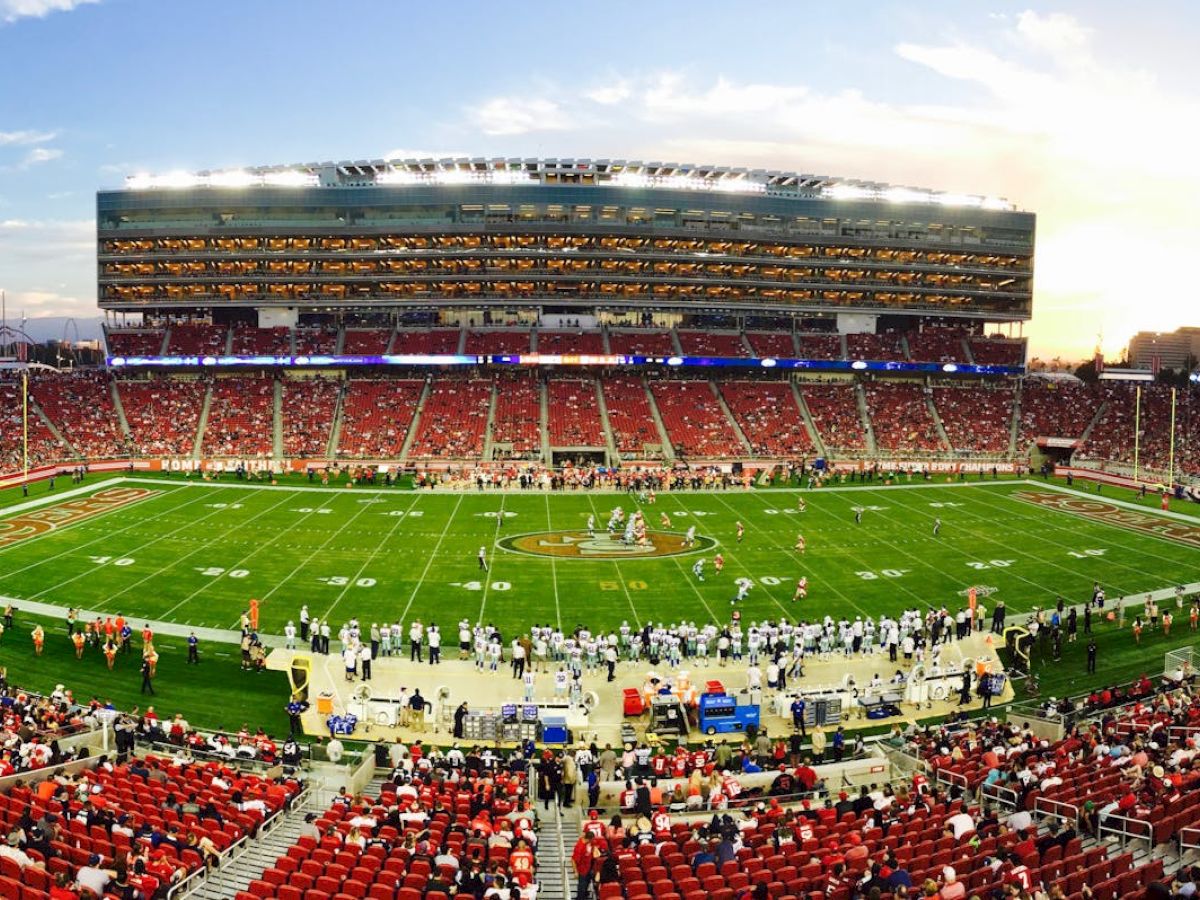 A large football stadium filled with spectators watching a game at sunset, with teams and staff on the field and bright lights overhead.