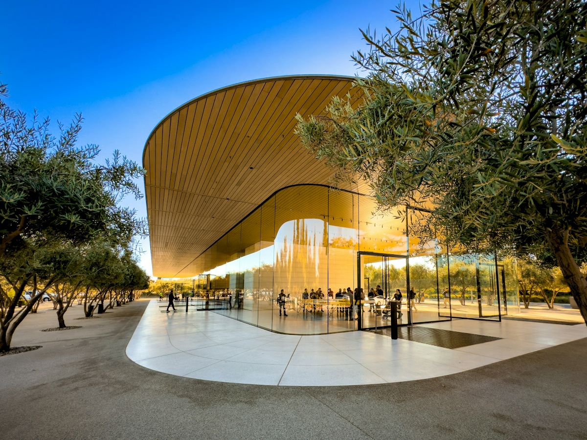 The image shows a modern building with curved glass walls and a wooden roof, surrounded by trees under a clear blue sky.