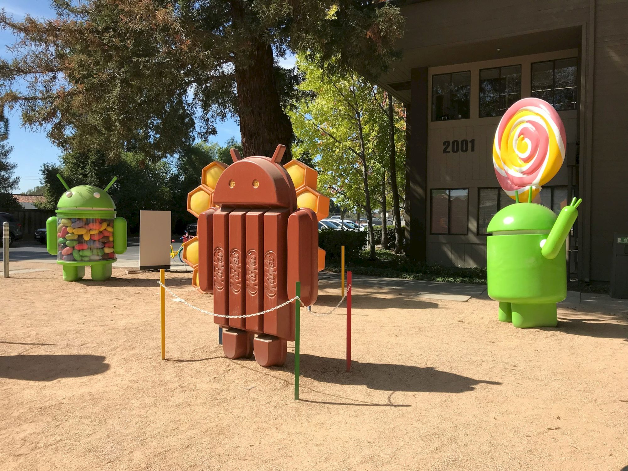 The image shows Android mascots themed as KitKat, Lollipop, and another version outdoors with trees and a building in the background.