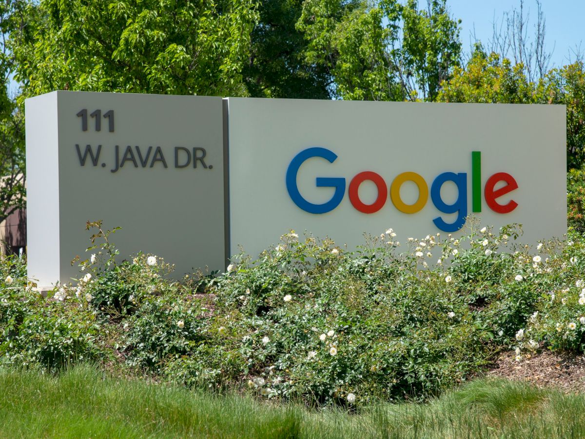 The image shows a Google sign at 111 W. Java Dr., surrounded by greenery and flowers.