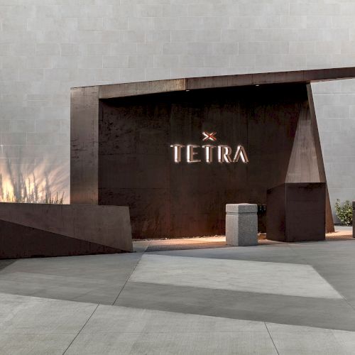 The image shows a modern architectural entrance with the word "TETRA" in glowing letters, featuring a fire feature on the left.