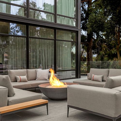 A modern outdoor seating area with a central fire pit, surrounded by sofas and large glass windows in the background.