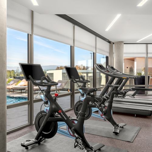 The image shows a modern gym with exercise bikes, treadmills, and a view of a pool outside large glass windows.
