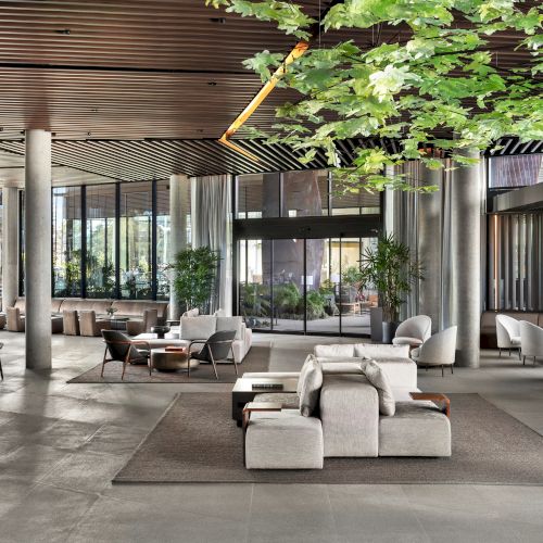 A spacious, modern lobby with seating areas, plants, and large glass windows. The ceiling features wooden panels and hanging leaves.