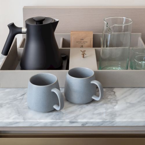 A black electric kettle, two gray mugs, a carafe, glasses, and instant coffee packets on a marble countertop.