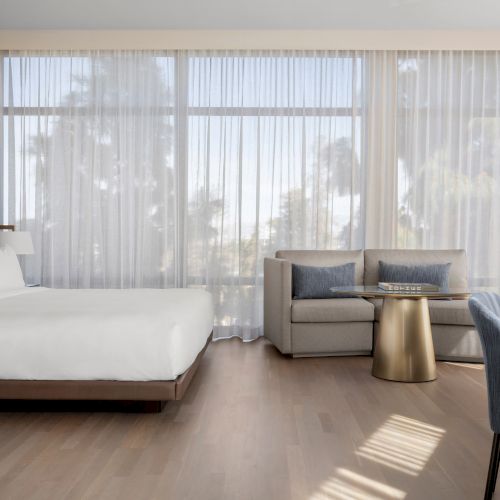 A modern hotel room with a bed, sofa, coffee table, and desk chair. Large windows with sheer curtains allow natural light in.
